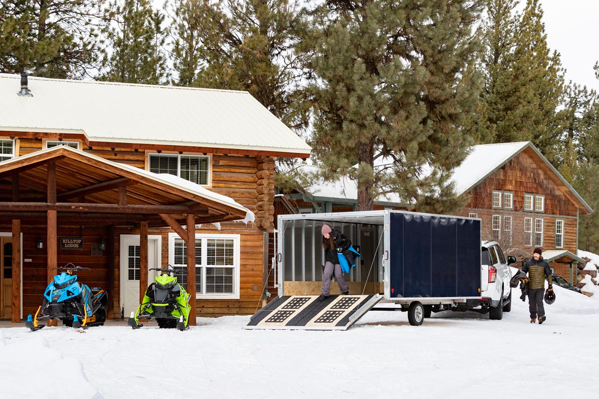 High Country Enclosed Crossover 2.0 Snow Trailer Parked In Front of Cabins With Snowmobiles